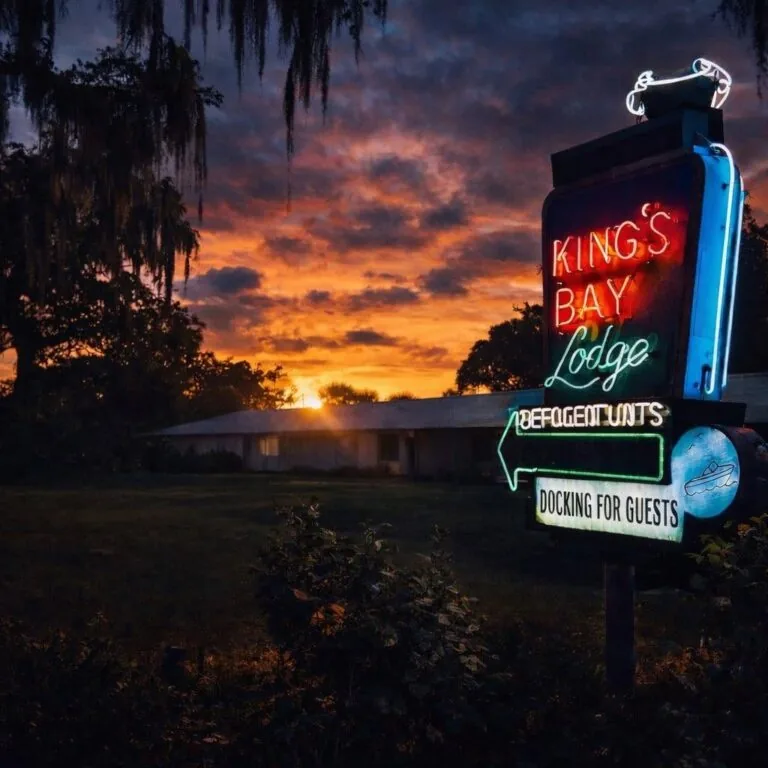 A glowing vintage neon sign for King's Bay Lodge at sunset with a dramatic orange sky and a motel building in the background.