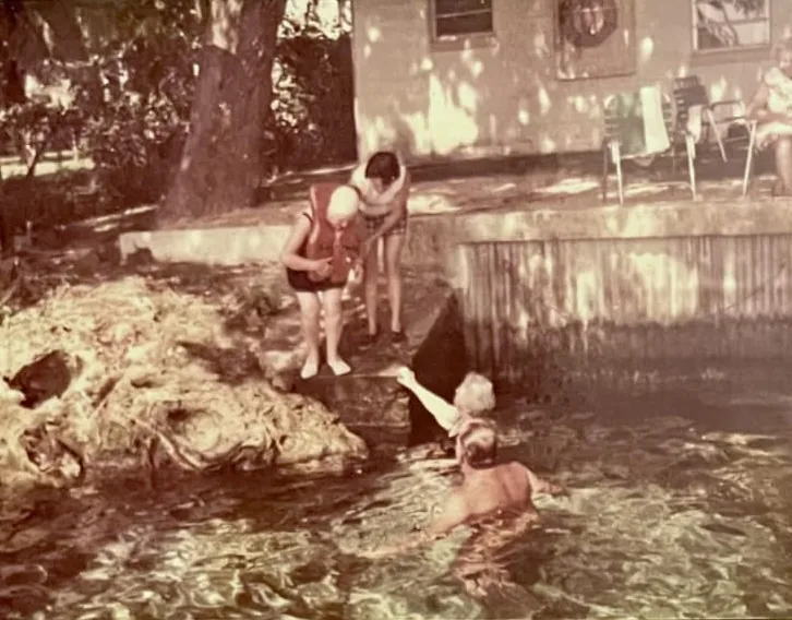 Vintage sepia photo of a family at the water's edge by a building, with a child in a life jacket and two people swimming.
